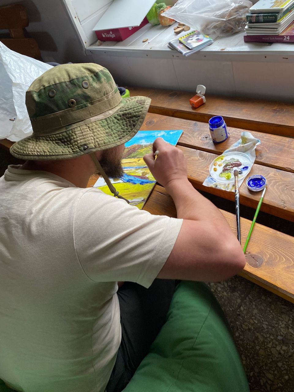 Учасник арт-терапії малює гірський пейзаж акриловими фарбами, сидячи за дерев’яним столом