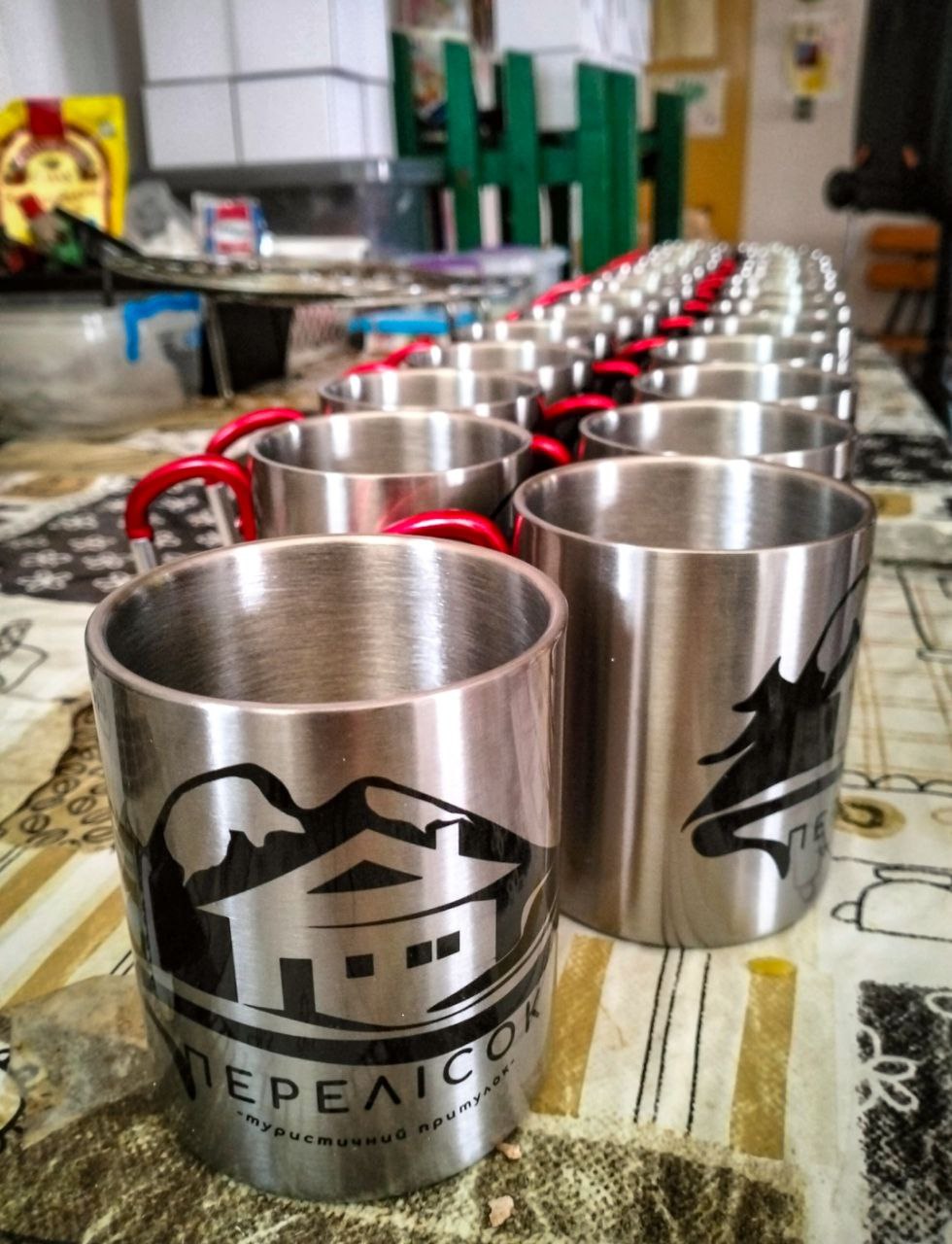 Металеві туристичні горнята з логотипом притулку «Перелісок» стоять у ряд на столі.