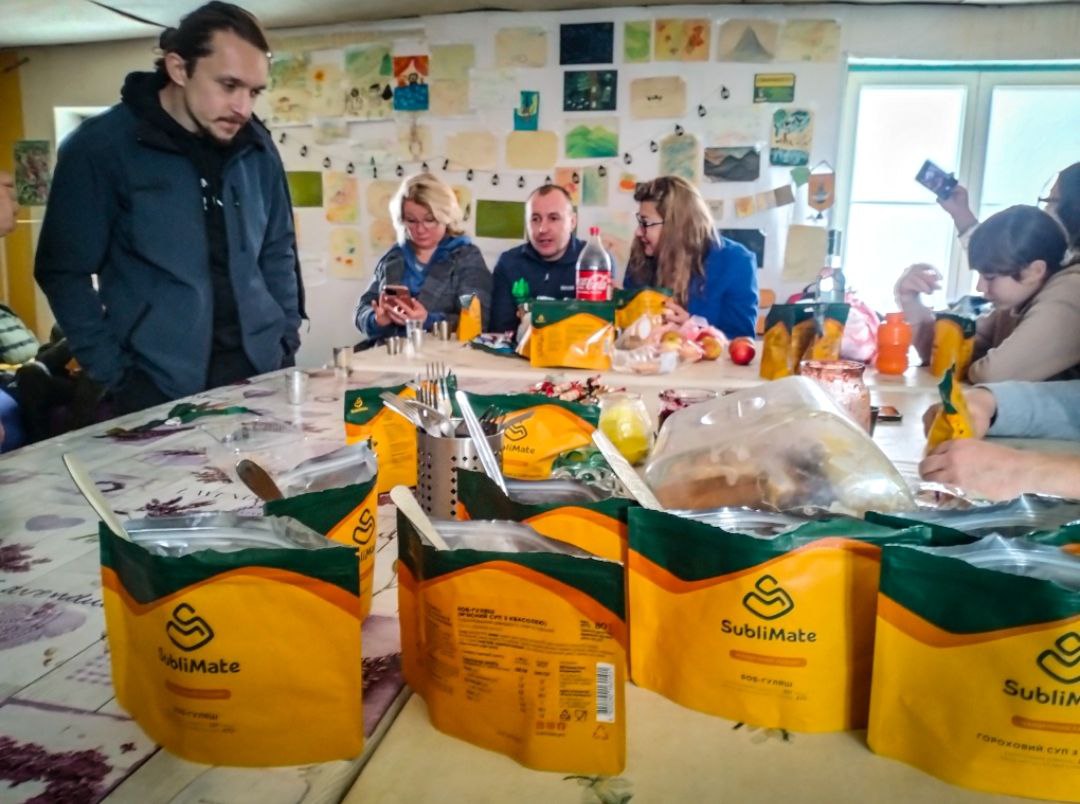 Група учасників у теплому приміщенні за великим столом. На столі — сублімовані страви для обіду.